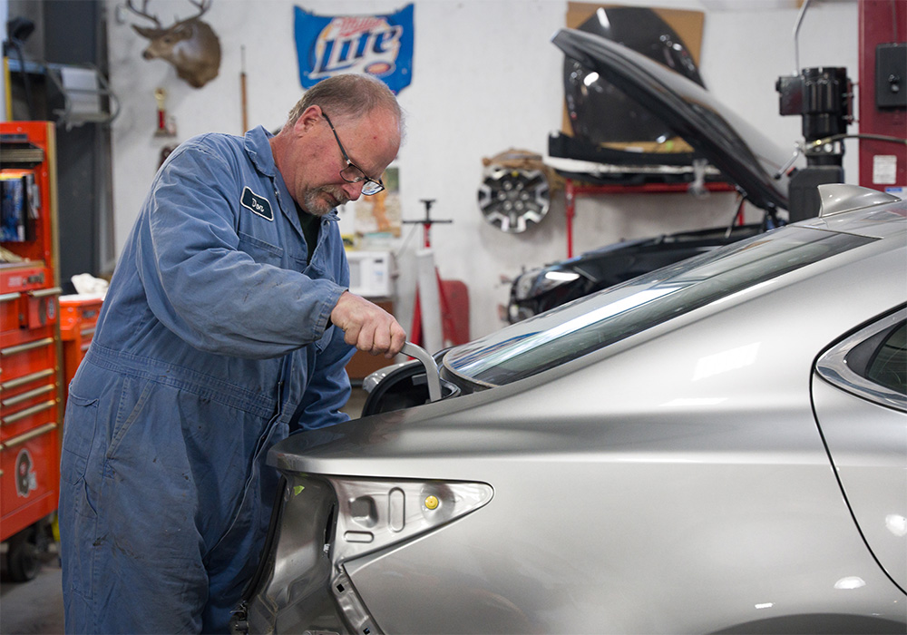 A person leans over the trunk of a car and inspects the hinge mechanism.
